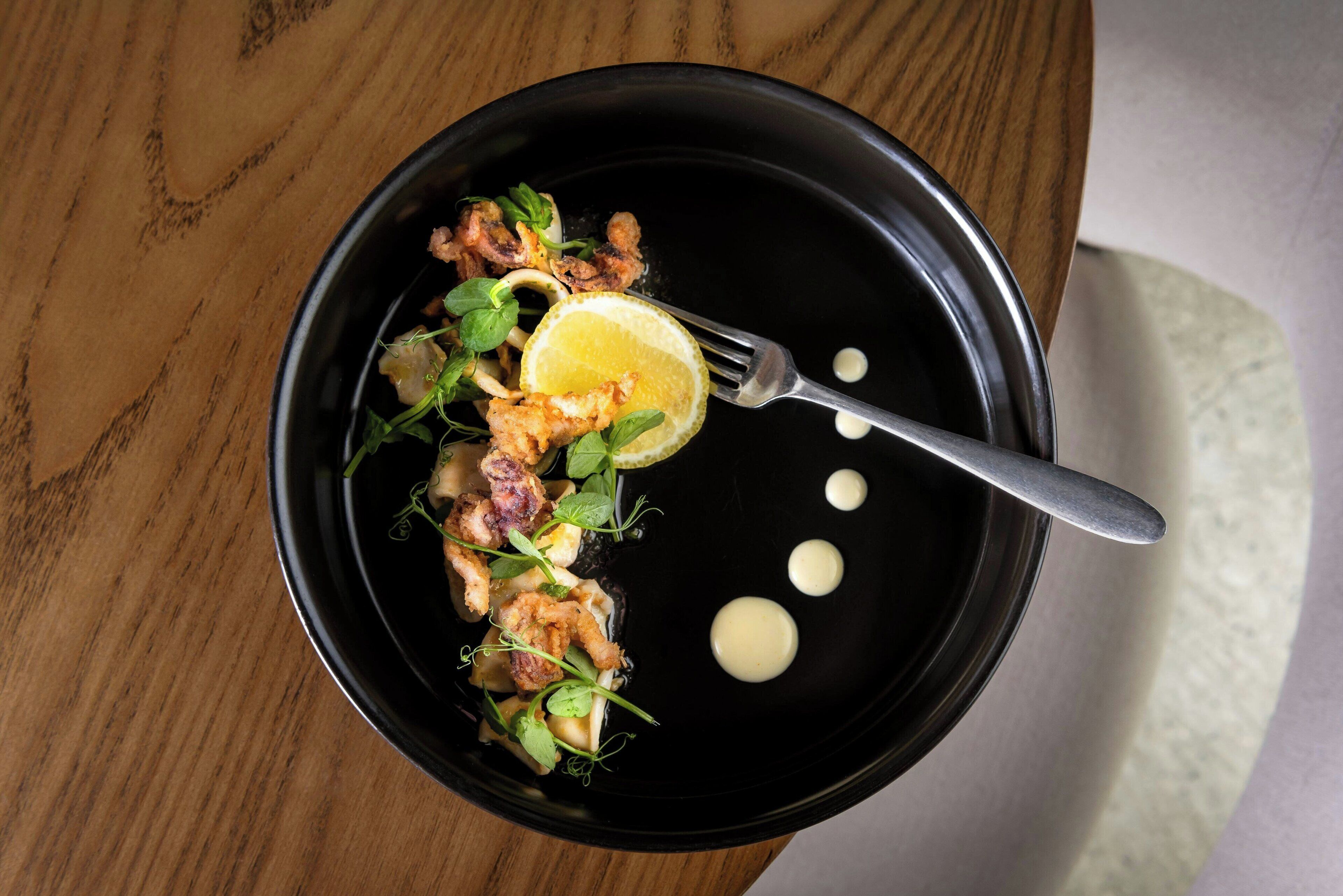 A black plate with food, a fork, and three dollops of sauce on a wooden table.