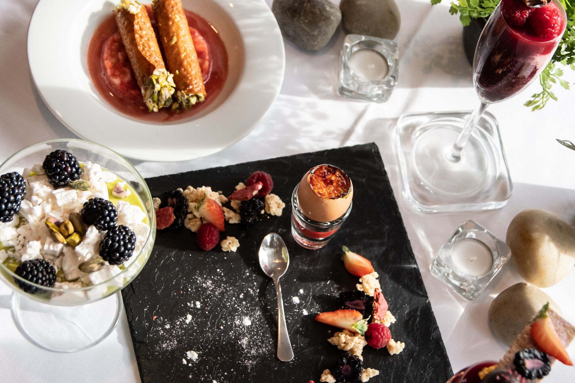 Una mesa de postres con diversos dulces, una copa de vino tinto y una vela sobre un mantel blanco.