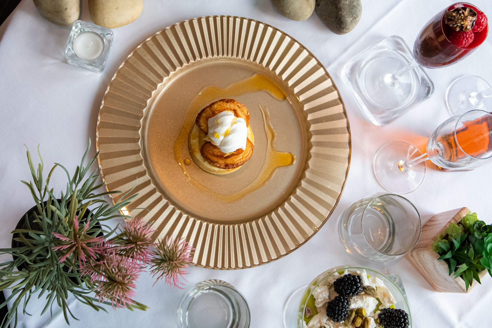 Un plato de postre con un pastel, almíbar y una vela pequeña sobre una mesa con copas de vino y un bol de frutos rojos.