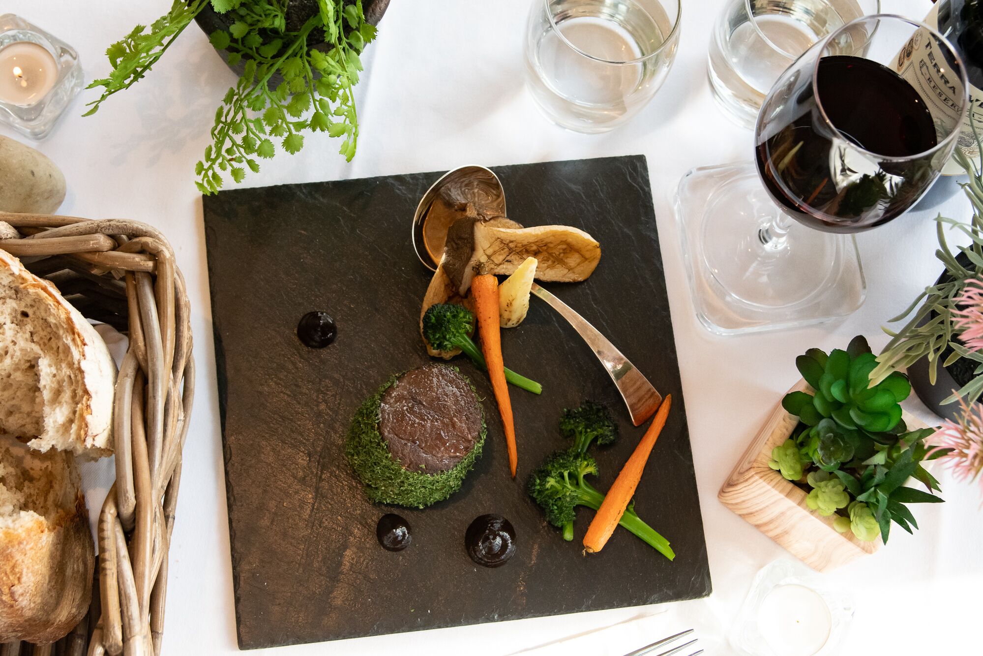 A dining table with a black slate board, various vegetables, a piece of meat, and a glass of red wine.