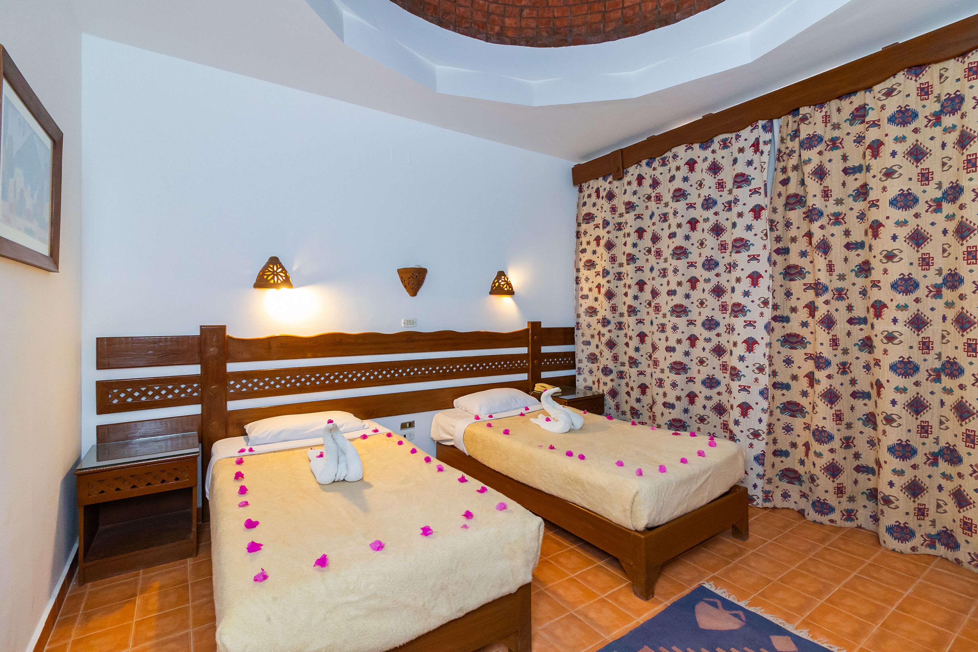 A hotel room with two beds, a wooden headboard, wall-mounted lamps, patterned curtains, and a tiled floor.