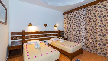 A hotel room with two beds, a wooden headboard, wall-mounted lamps, patterned curtains, and a tiled floor.