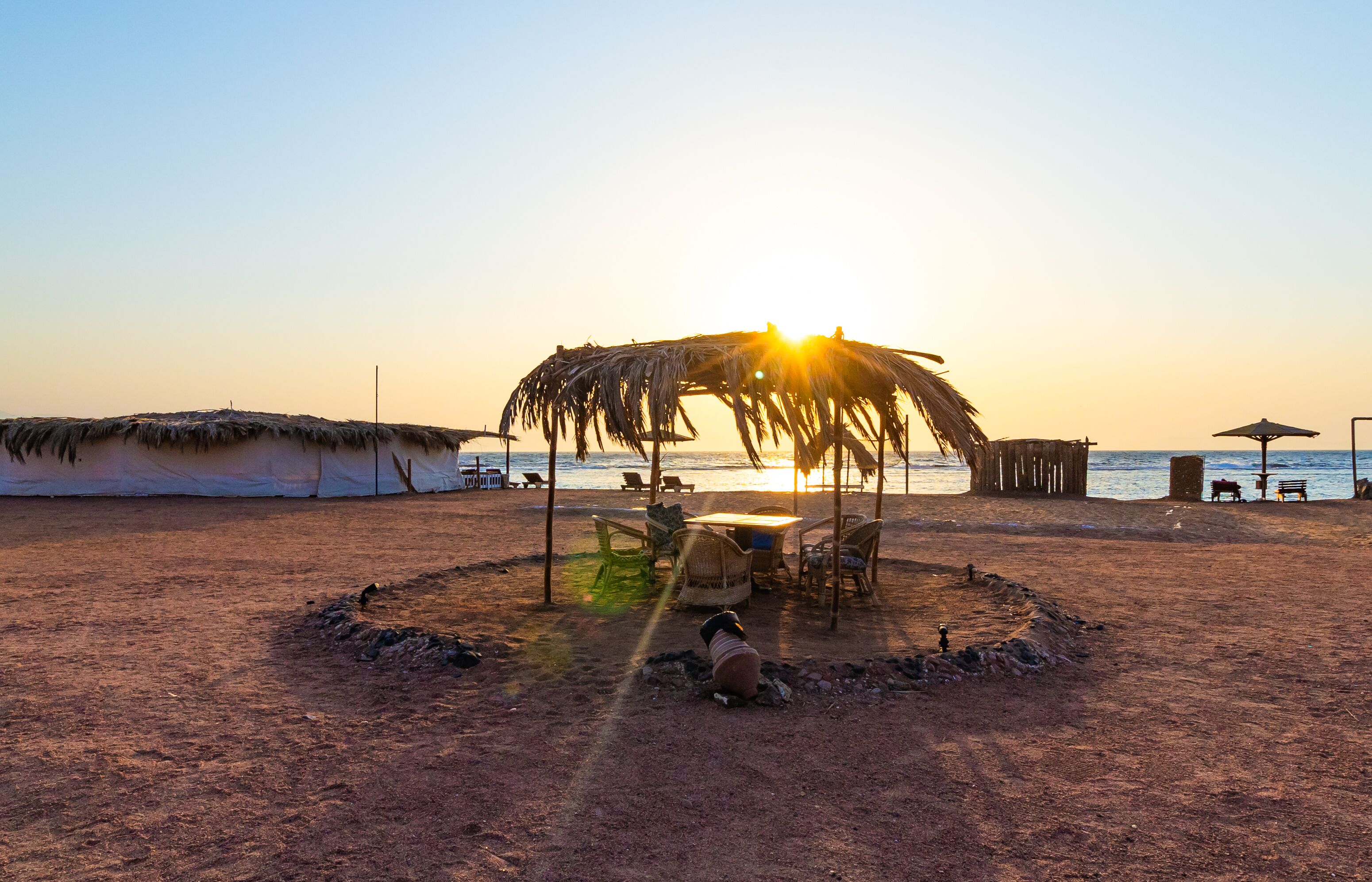 A beach with a thatched umbrella and chairs, a small fire pit, and a view of the sea at sunset.