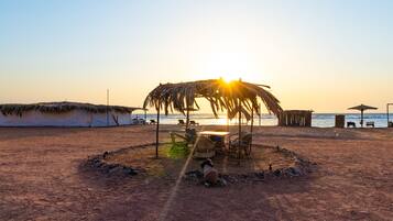 A beach with a thatched umbrella and chairs, a small fire pit, and a view of the sea at sunset.