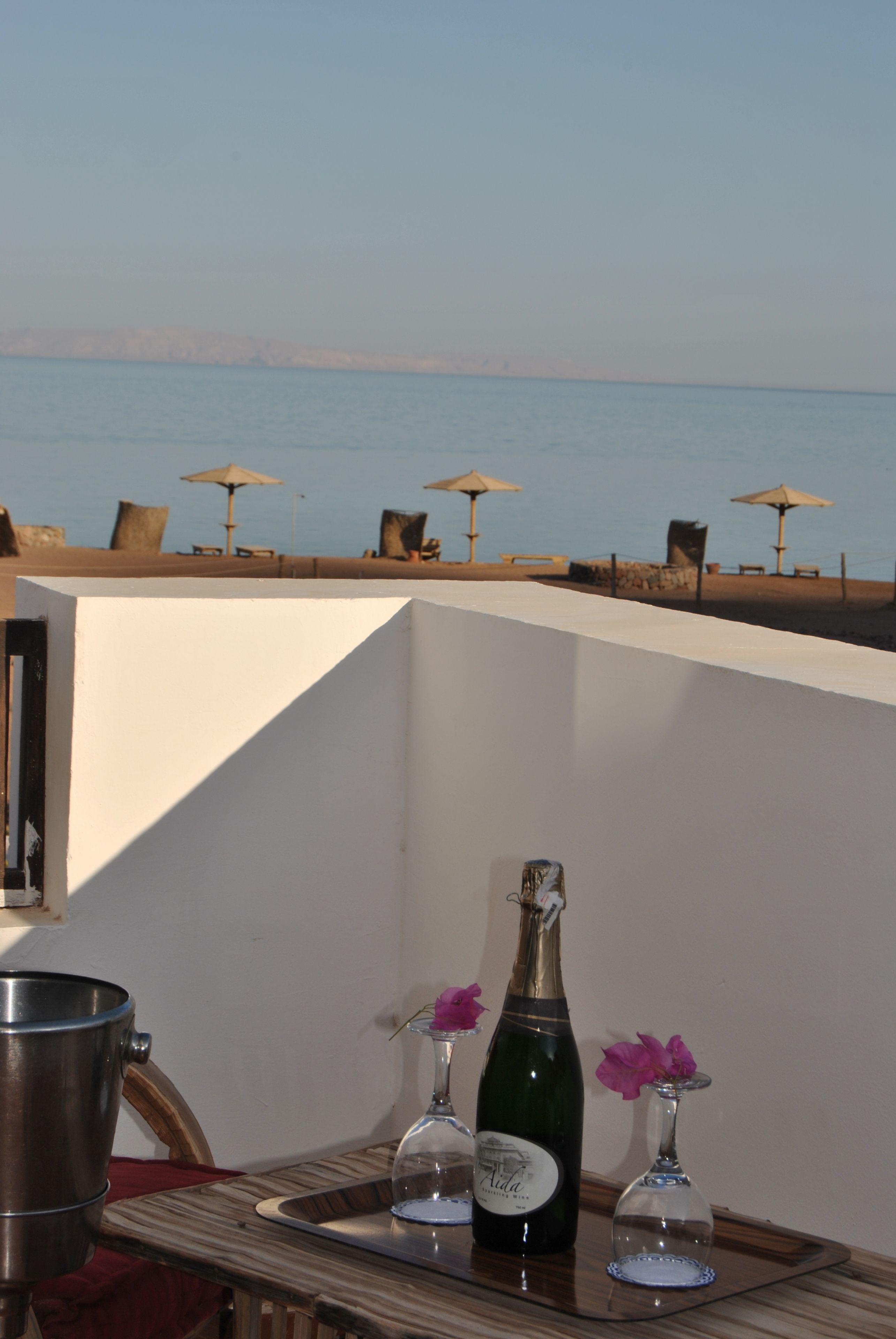 Um terraço com vista para o mar, com uma mesa, uma garrafa, copos e cadeiras de praia com guarda-sóis.