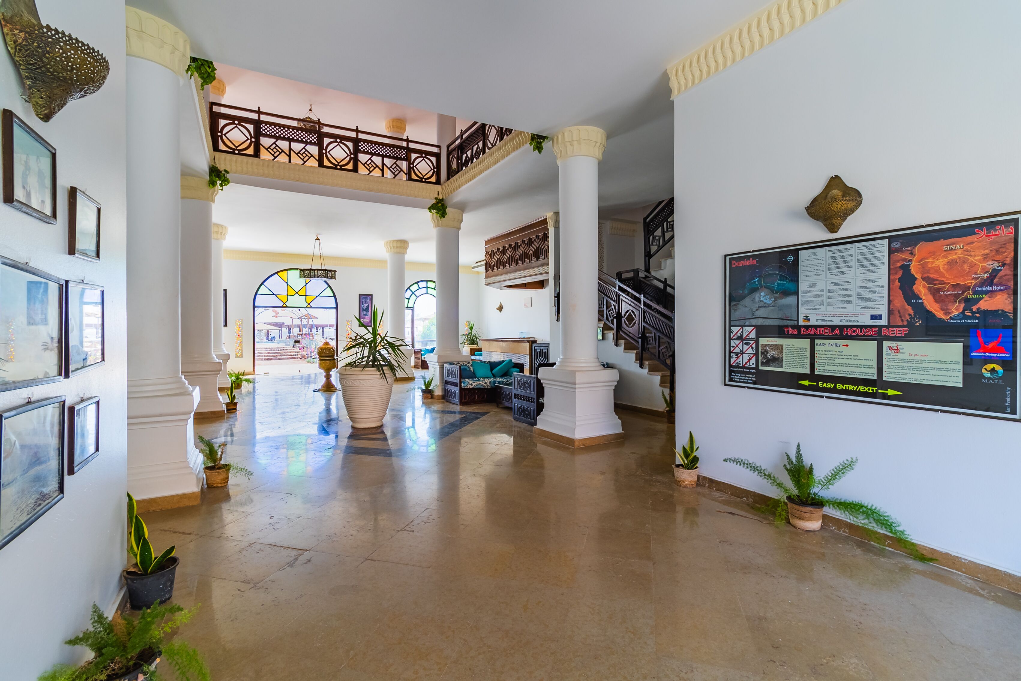 Un'ampia hall con soffitto alto, colonne e una scalinata. Sono presenti piante in vaso e quadri alle pareti.