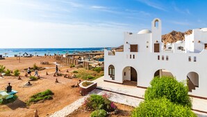 A white building with arches and a tower, situated near a beach with people walking and umbrellas. - Daniela Village Dahab (Dahab)