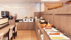 Un espace buffet avec différents postes de service proposant divers plats, notamment une machine à café, ainsi qu’un coin salon avec des tables et des chaises en bois.