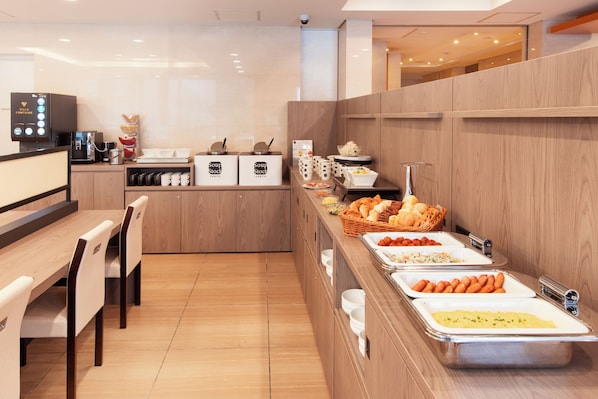Zona de buffet con diferentes estaciones de comida, incluyendo una máquina de café, y un área de estar con mesas y sillas de madera.