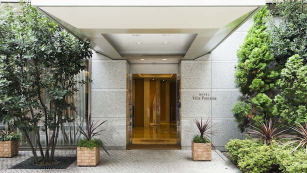 L’entrée d’un hôtel, dotée d’un porche abrité, d’une enseigne indiquant « Hôtel Villa Fontaine » et de plantes en pot disposées autour de l’entrée.