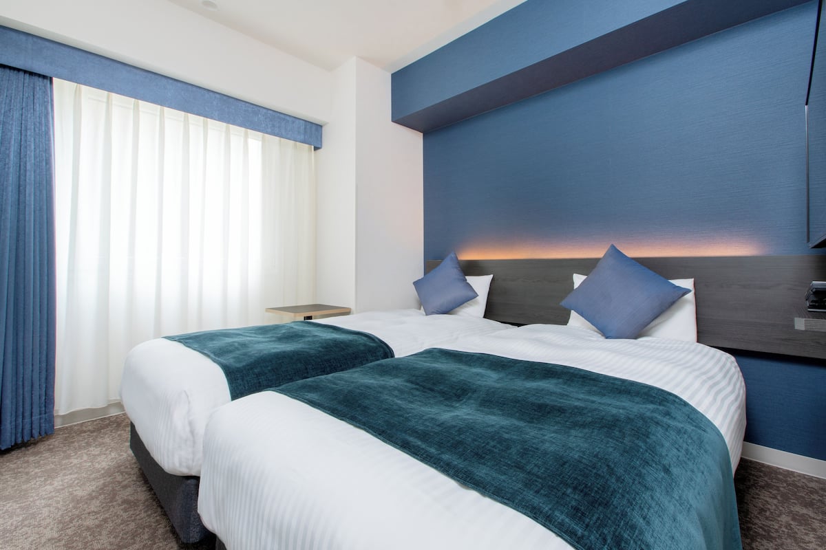 Habitación de hotel con dos camas, una mesita de noche y un ventanal con cortinas.
