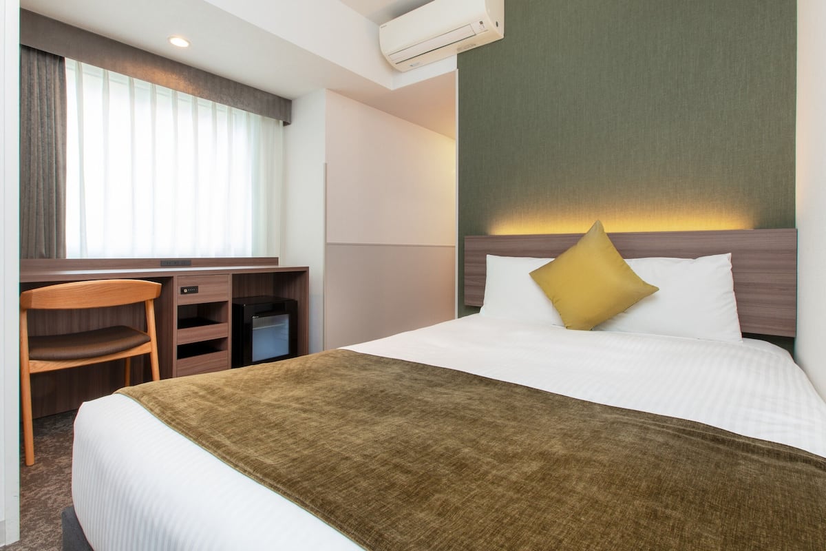 Una habitación de hotel con cama, escritorio, silla, aire acondicionado y ventana con cortinas.