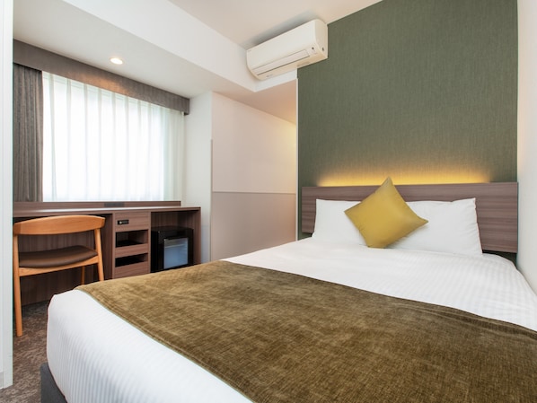Una habitación de hotel con cama, escritorio, silla, aire acondicionado y ventana con cortinas.