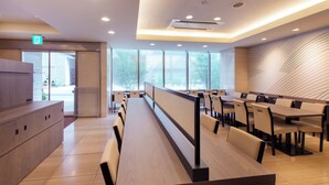 Un espace repas moderne avec de longues tables en bois et des chaises beiges, de grandes fenêtres et un mur aux lignes courbes.