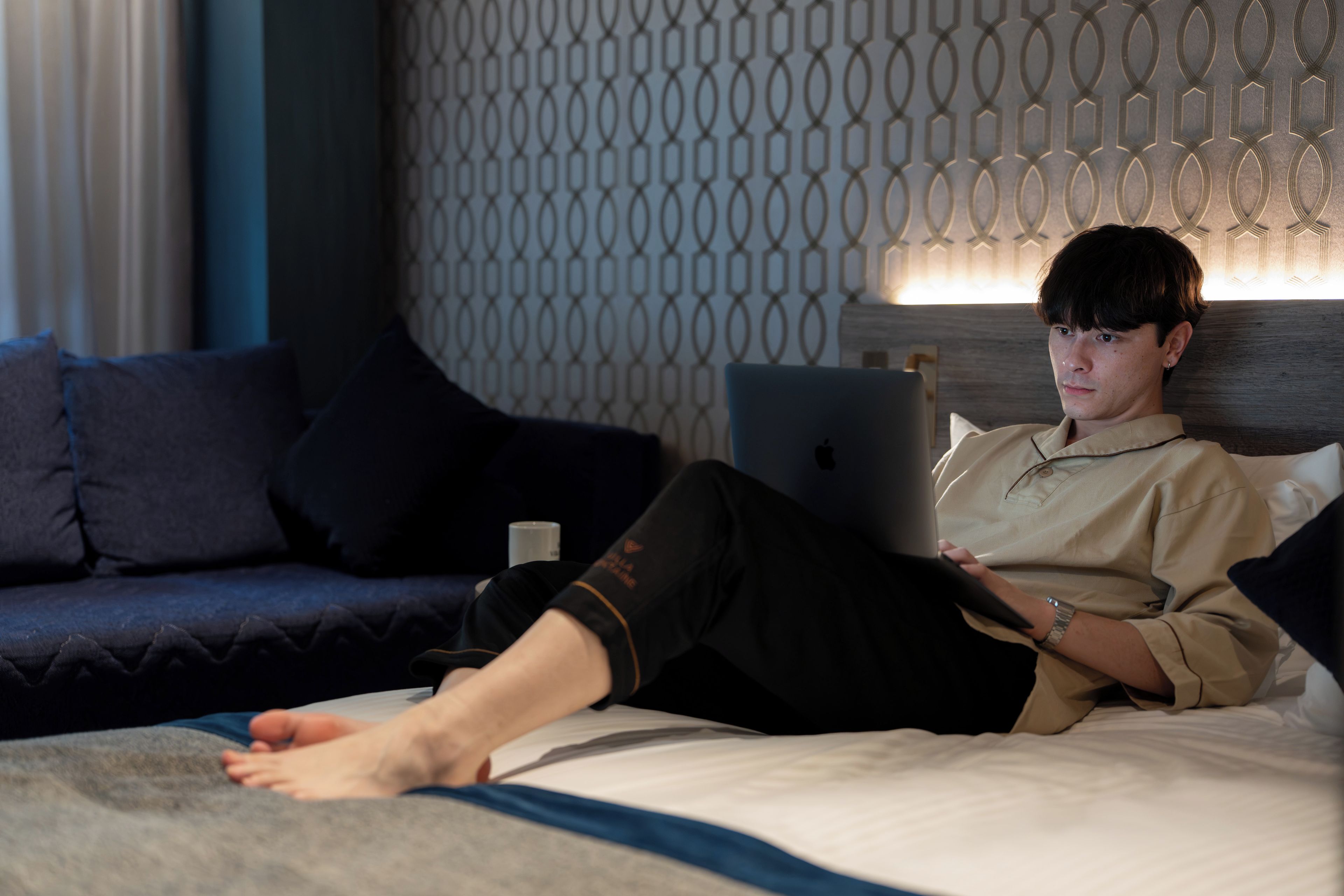 A person sitting on a bed with a laptop, a blue couch, and patterned wallpaper in a room.