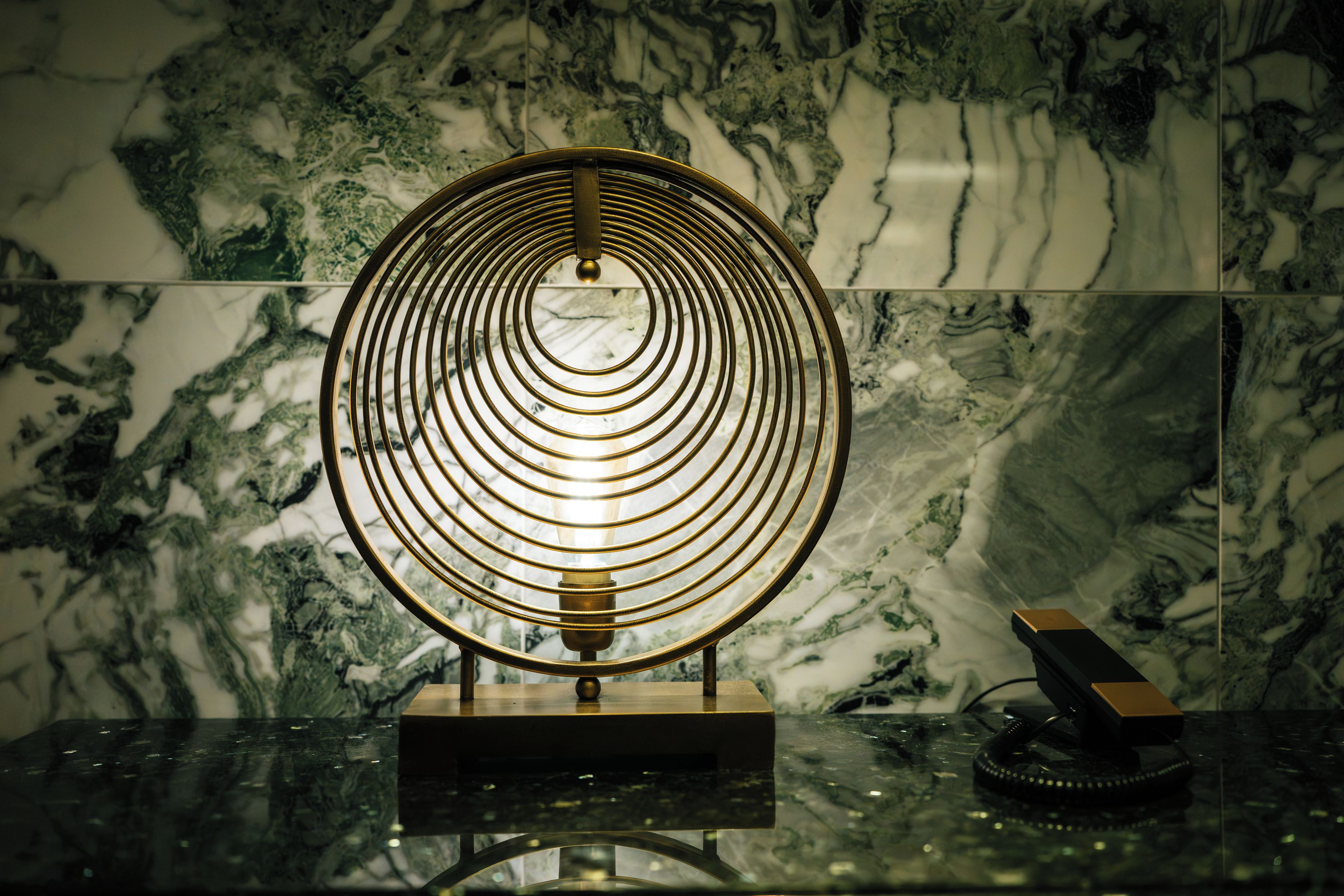 A circular metallic object with concentric rings on a reflective surface, possibly a decorative piece or a functional item, against a marble background.