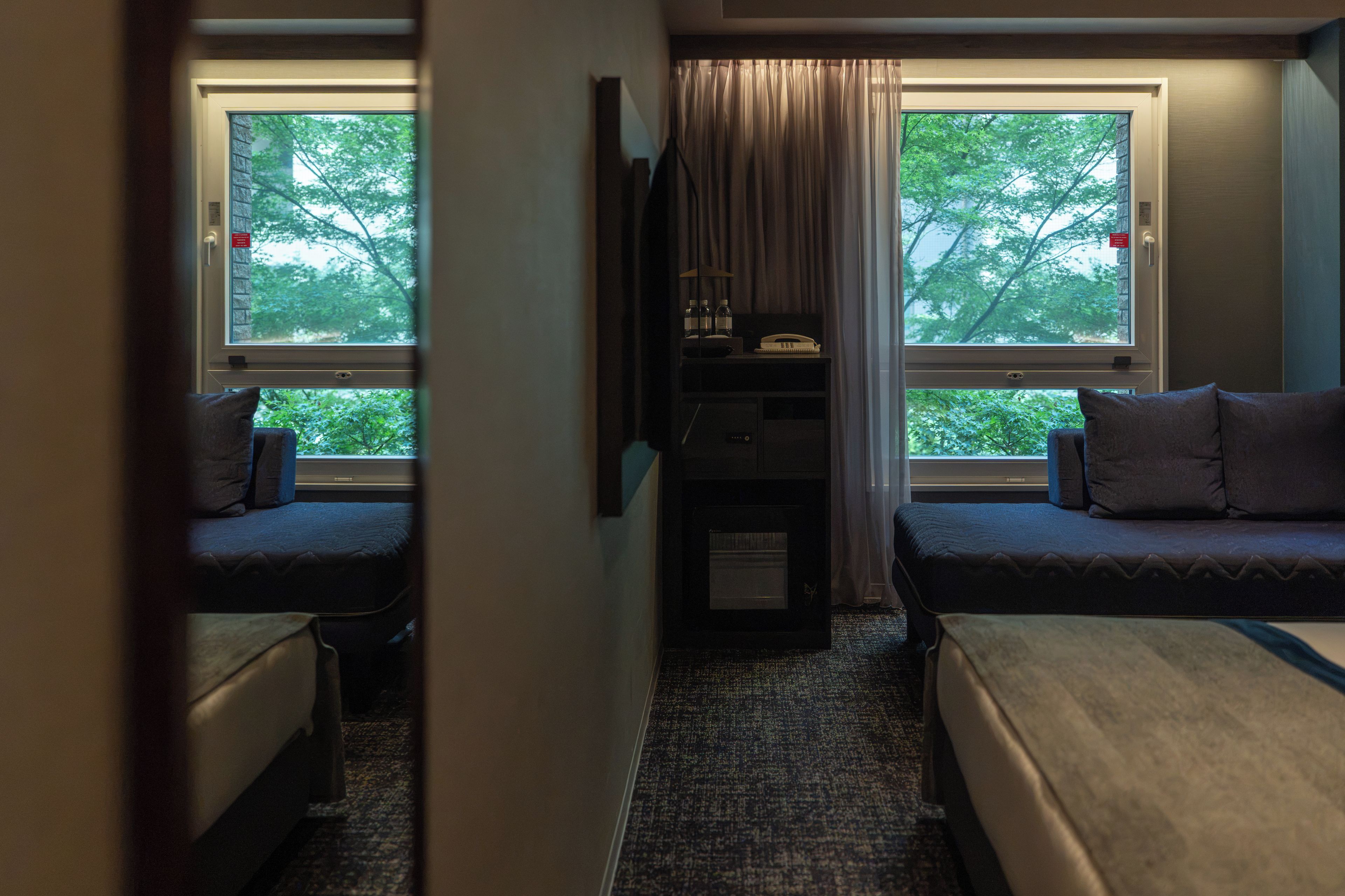Une chambre d’hôtel de taille modeste, équipée de deux lits simples, d’un petit bureau avec un téléphone et offrant une vue sur les arbres par les fenêtres.