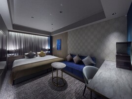 A modern hotel room with a large bed, a sofa, a small round table, and a desk with a chair.