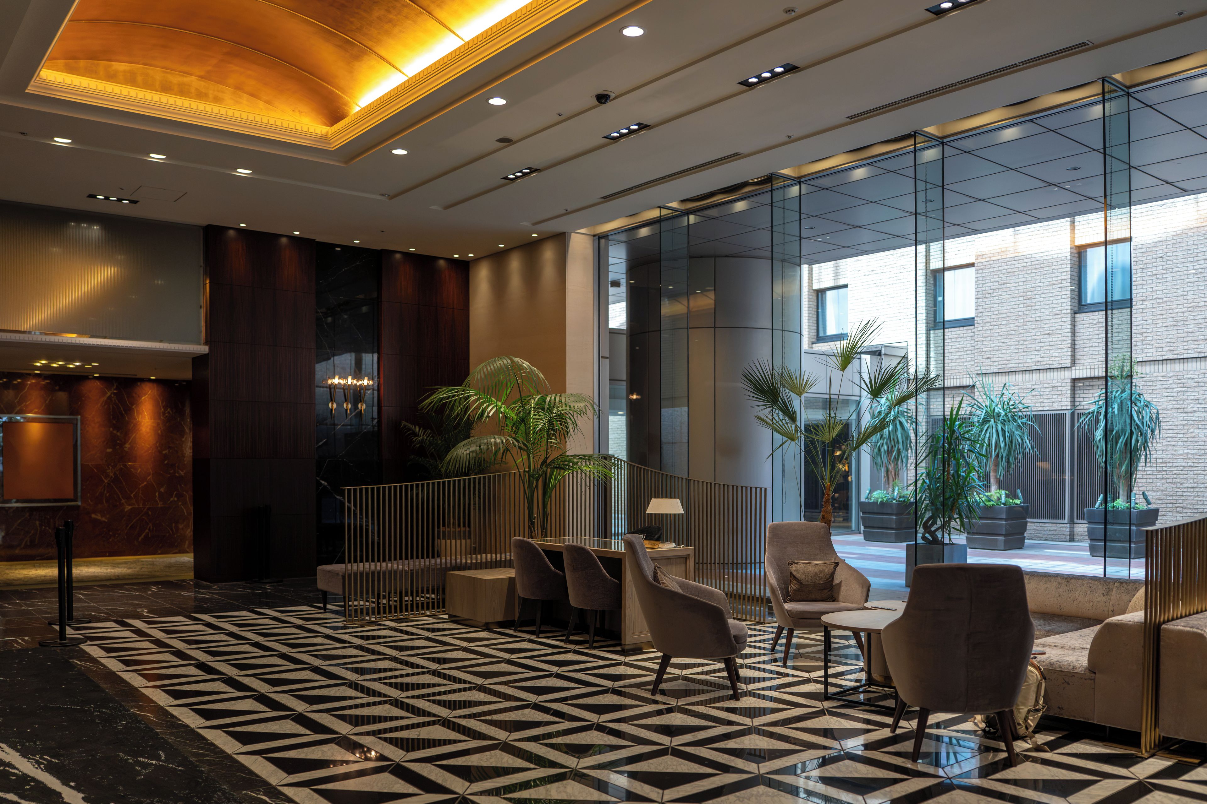 A lobby with a geometric patterned floor, seating area with chairs and a sofa, a reception desk, and a glass partition with a view of the outside.