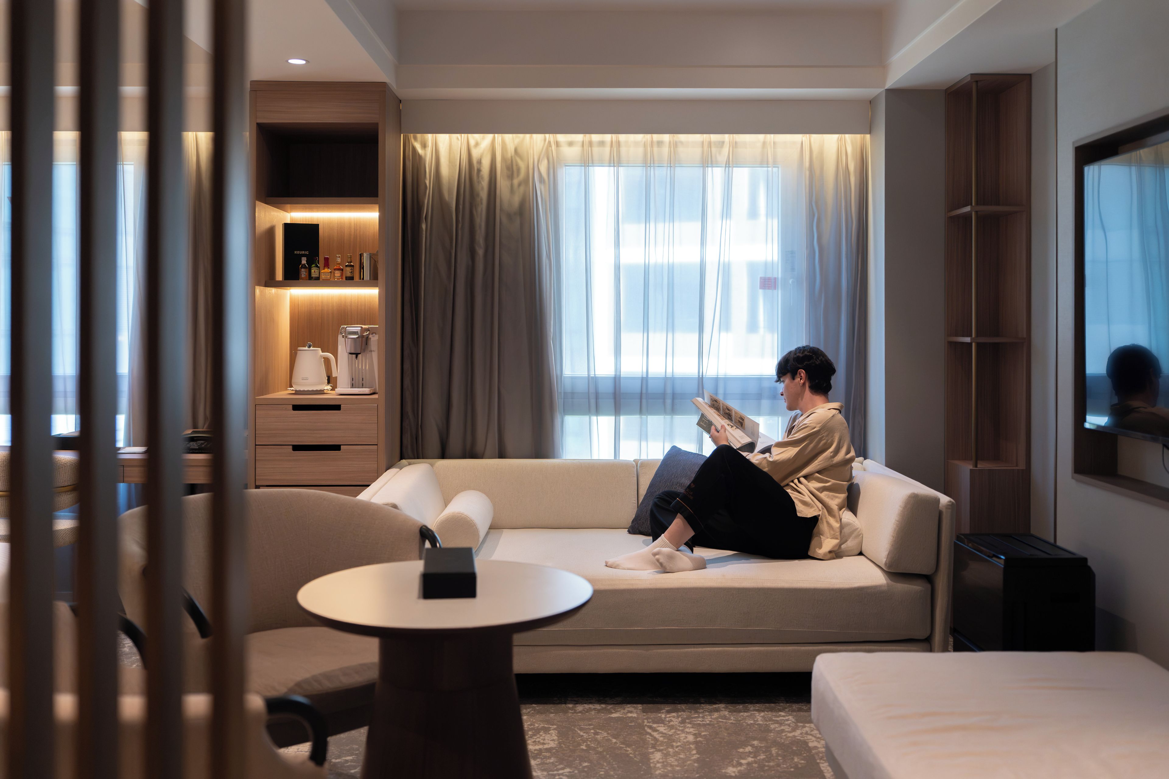 A modern hotel room with a sofa, a small table, a coffee machine, and a TV.