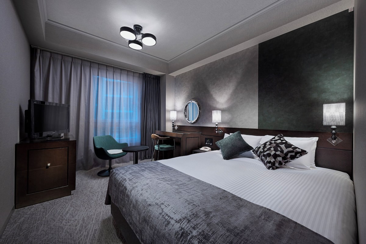 Une chambre d’hôtel moderne dotée d’un grand lit, d’un bureau avec une chaise, d’une télévision et d’une grande fenêtre avec des rideaux.