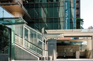 A modern hotel entrance with a glass facade, a sign for Hotel Villa Fontaine, and an adjacent shop and restaurant.