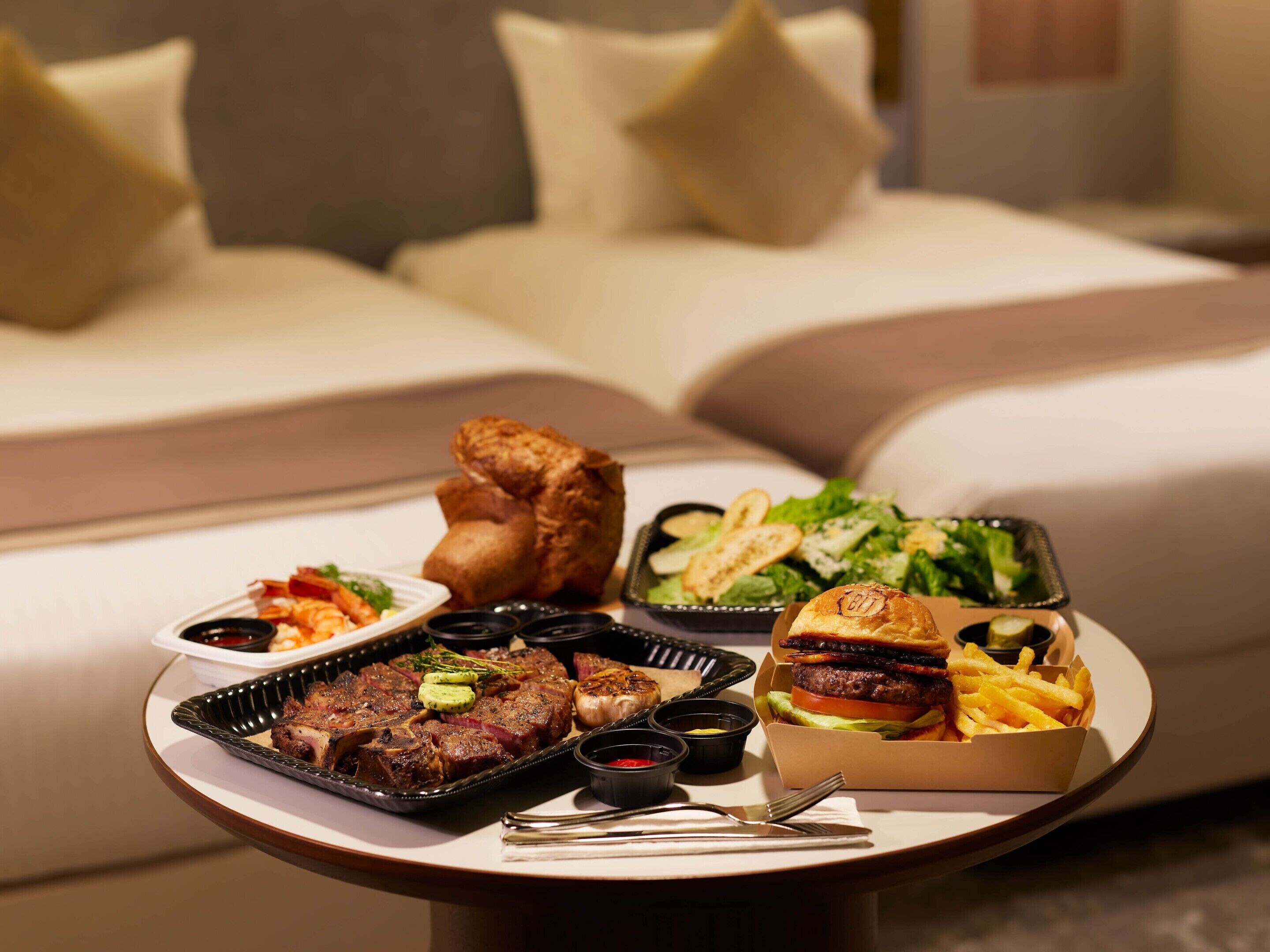 A dining table with a variety of dishes including steak, fries, and a burger, set in a room with a sofa and pillows in the background.