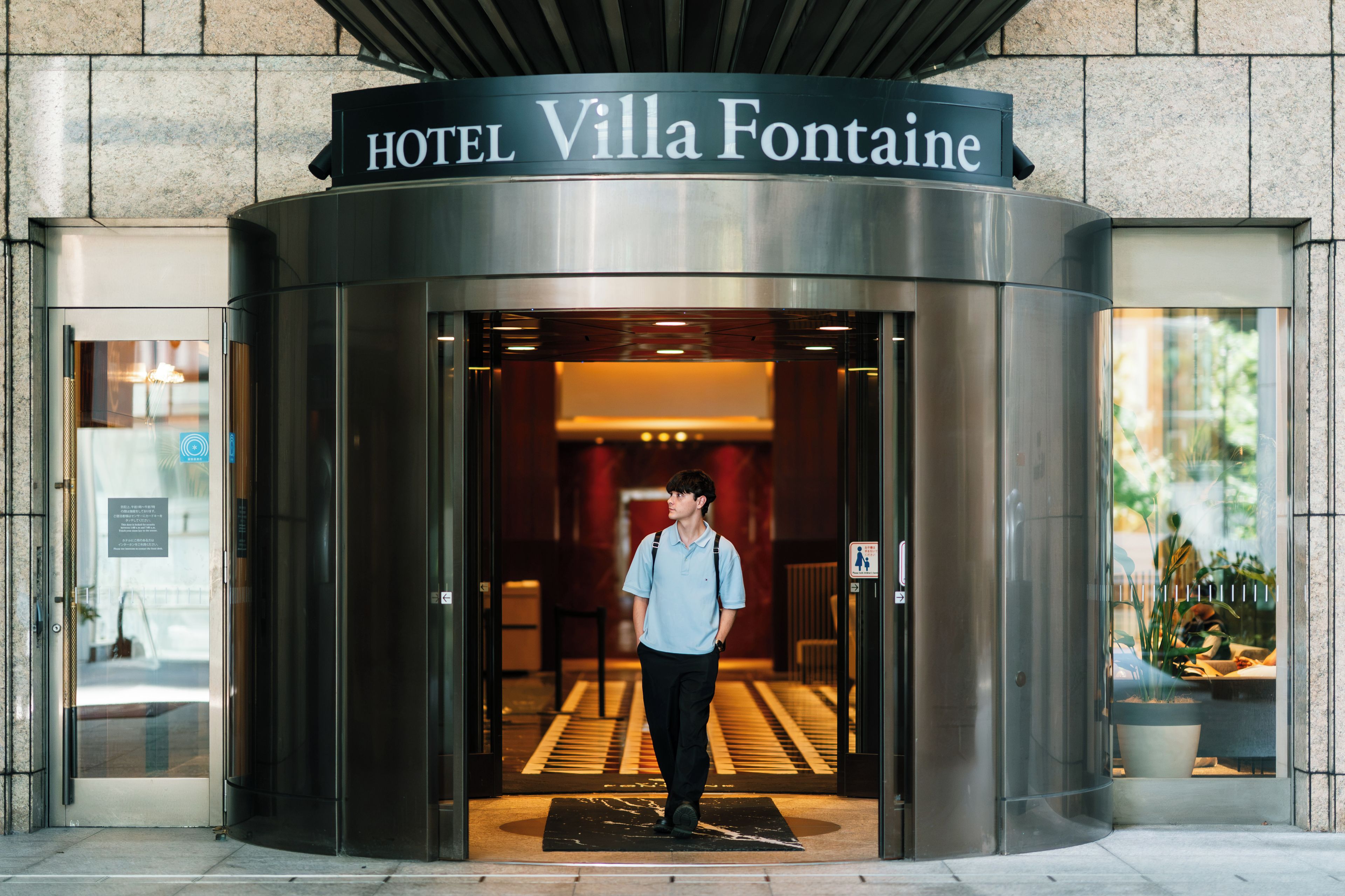 Entrada de un hotel con puerta giratoria, un cartel que dice 'HOTEL Villa Fontaine' y una persona de pie en el umbral.