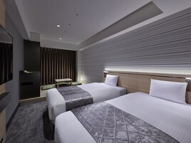 A modern hotel room with two beds, a flat-screen TV, a desk, and a large window with blinds.