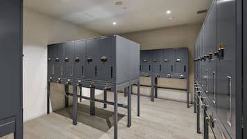 A row of grey lockers with metal handles and yellow locks, mounted on metal stands, in a room with beige walls and a speckled floor.