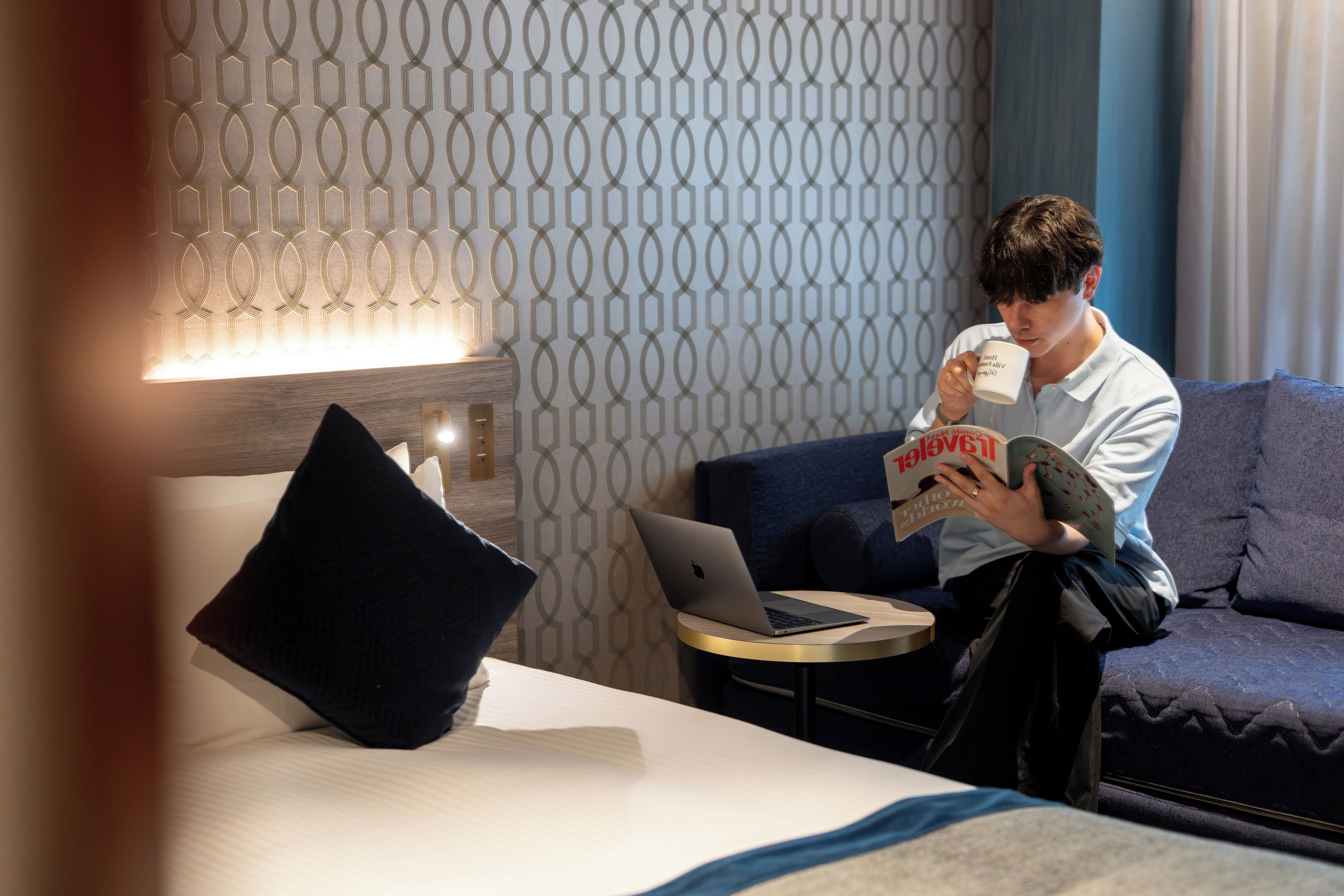 A person sitting on a couch with a laptop and a book, holding a mug.