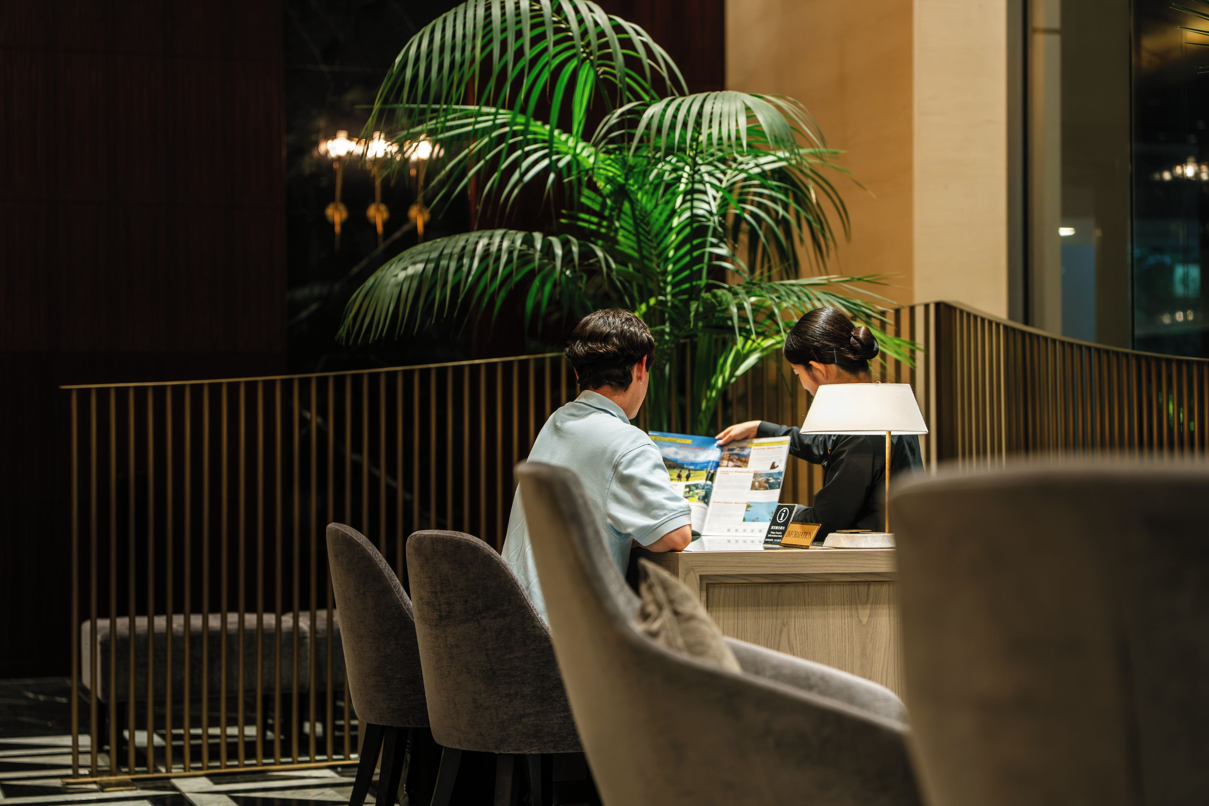 A reception desk with a large potted plant, a man and a woman examining a brochure, and a modern interior design with a railing and a lamp.