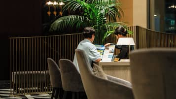 Un comptoir d’accueil agrémenté d’une grande plante en pot, un homme et une femme examinant une brochure, et un aménagement intérieur moderne avec une rambarde et une lampe.