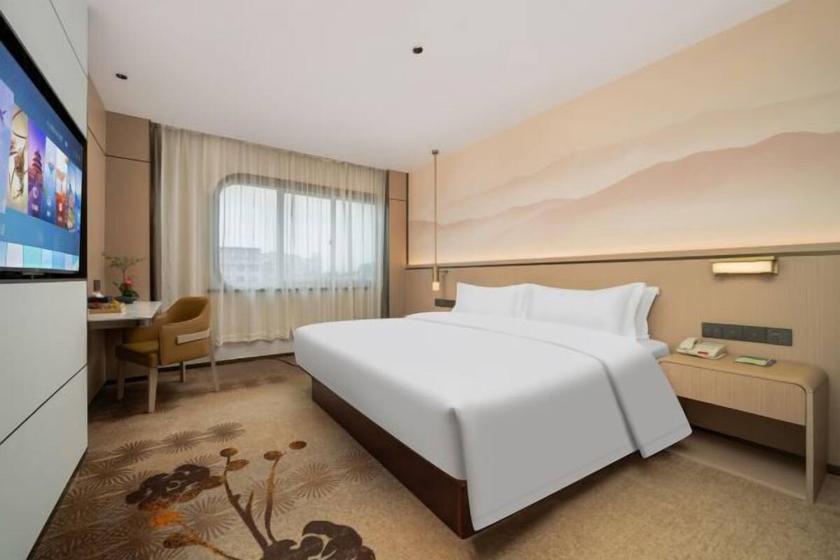 Ein Hotelzimmer mit Bett, Schreibtisch, Stuhl, Fernseher und einem großen Fenster mit Vorhängen.
