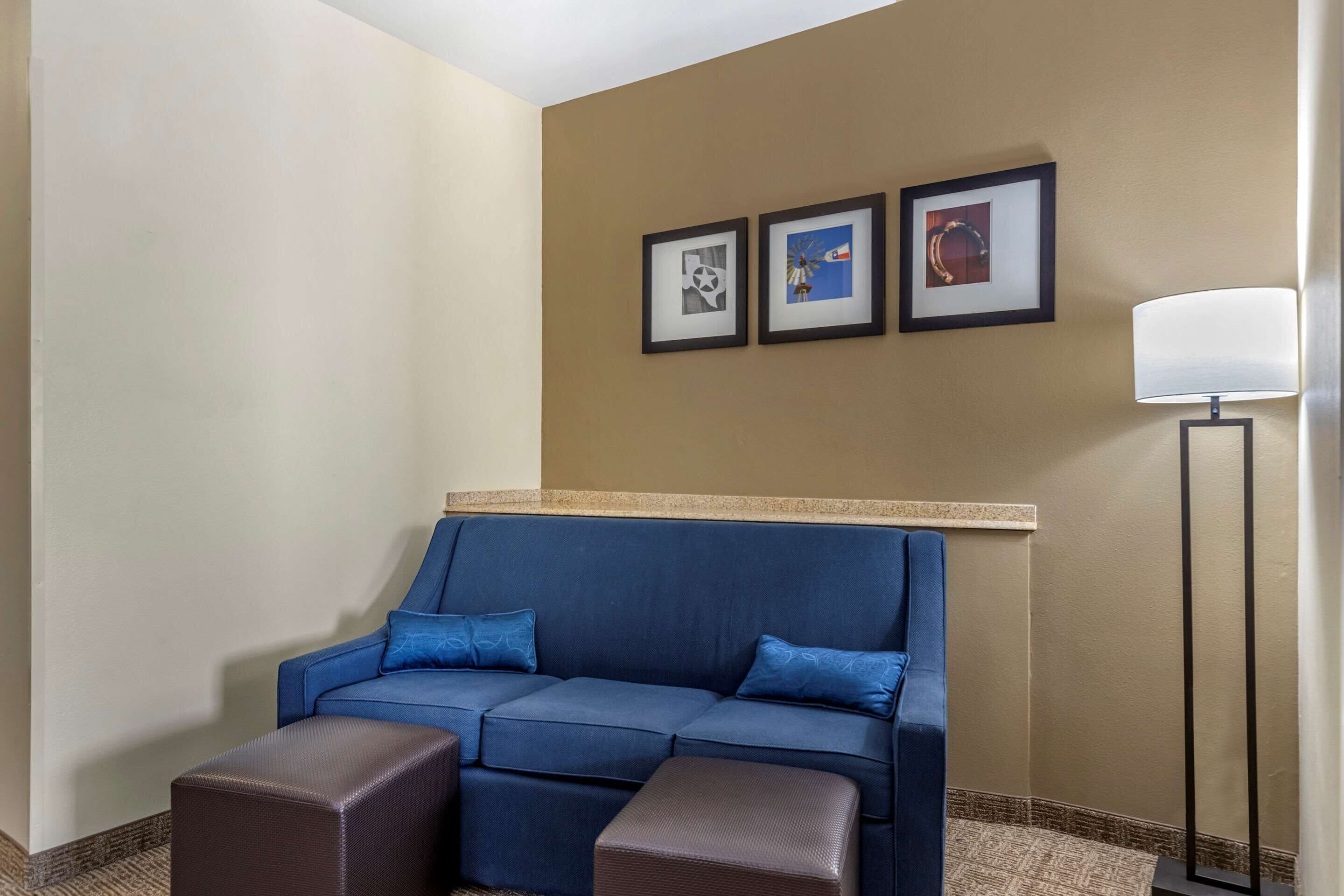 A hotel room with a blue sofa, two matching ottomans, a floor lamp, and three framed pictures on the wall.
