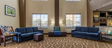 A spacious lobby with high ceilings, large windows, and a patterned carpet. There are two blue sofas and a small table with a lamp.
