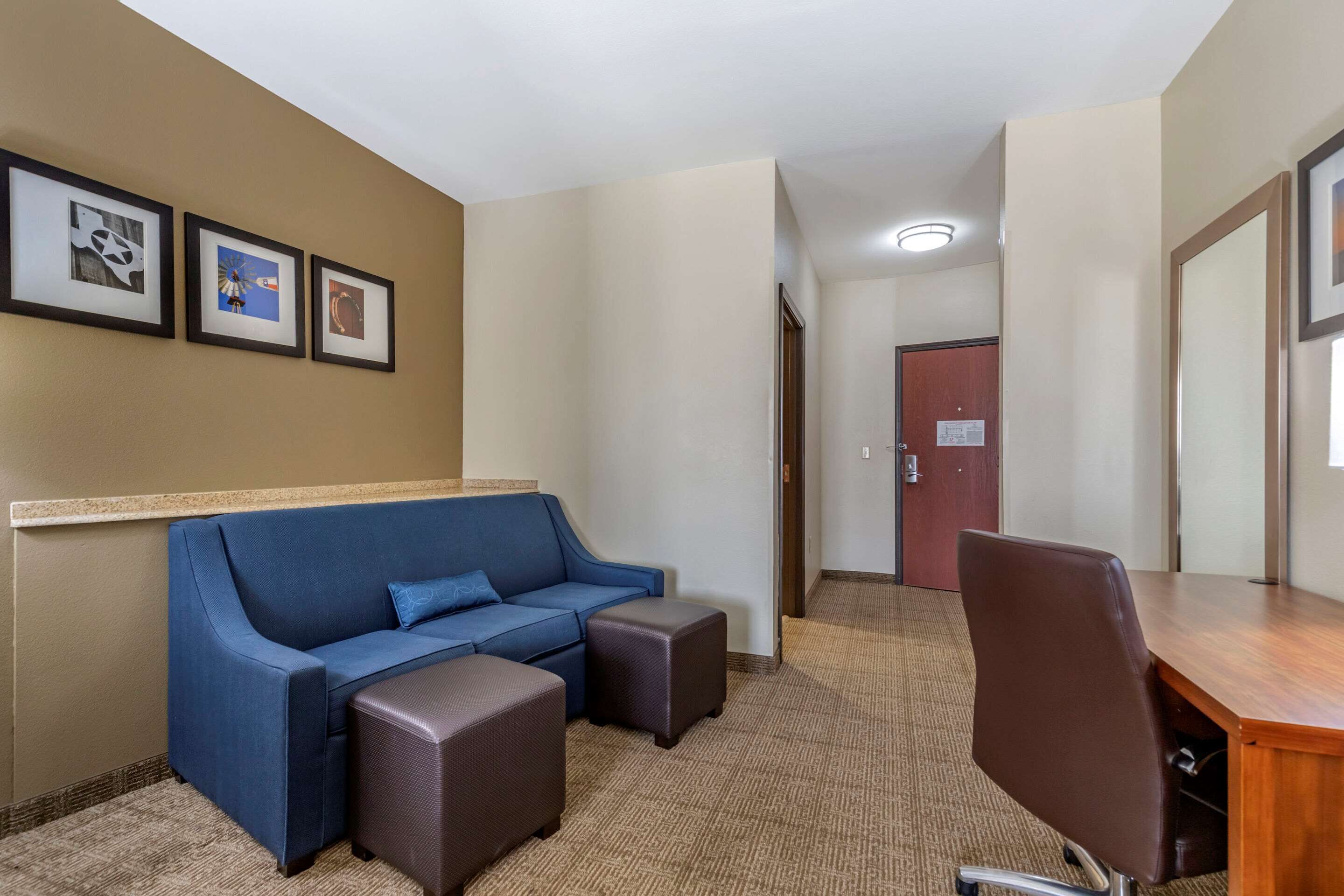 Una camera d'albergo con un divano blu, due poggiapiedi, una scrivania in legno e una sedia. Alle pareti si notano quadri incorniciati e, sullo sfondo, una porta.
