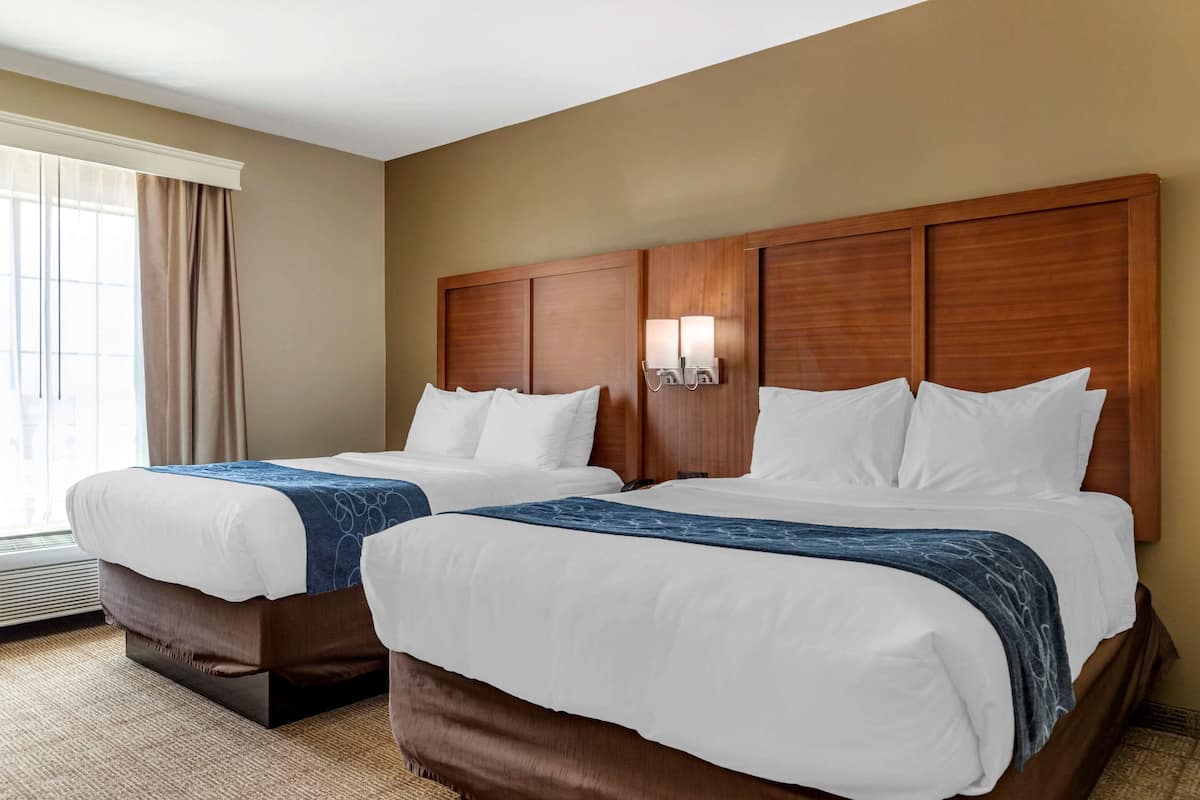 Quarto de hotel com duas camas, uma janela grande com cortinas e luminária fixada na parede.