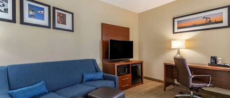 A hotel room with a blue sofa, a television on a wooden cabinet, a desk with a lamp, and framed pictures on the wall.