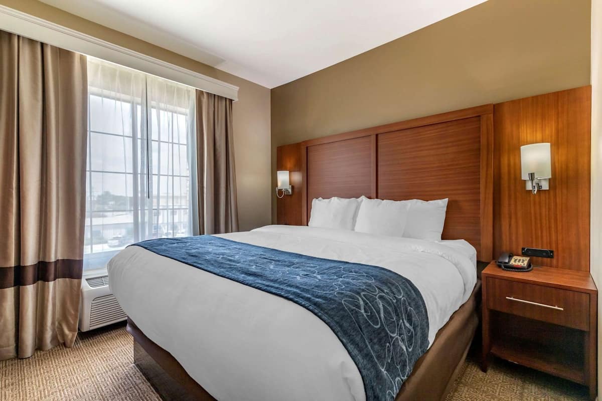 Quarto de hotel com uma cama grande, cabeceira de madeira, mesa de cabeceira com telefone e janela com cortinas.