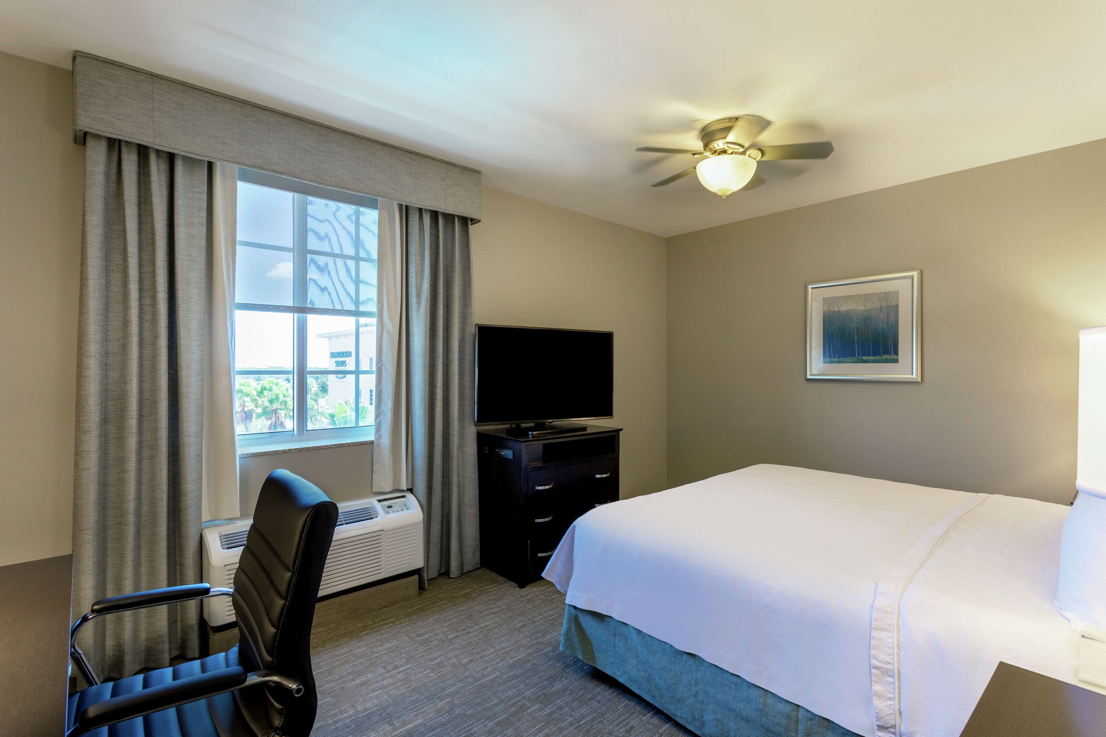 Quarto de hotel com uma cama, uma escrivaninha com cadeira, televisão e ventoinha no teto.