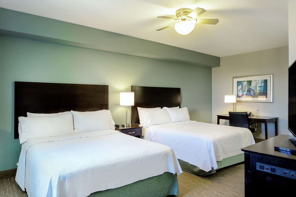 A hotel room with two beds, a desk, a chair, a ceiling fan, and a painting on the wall.