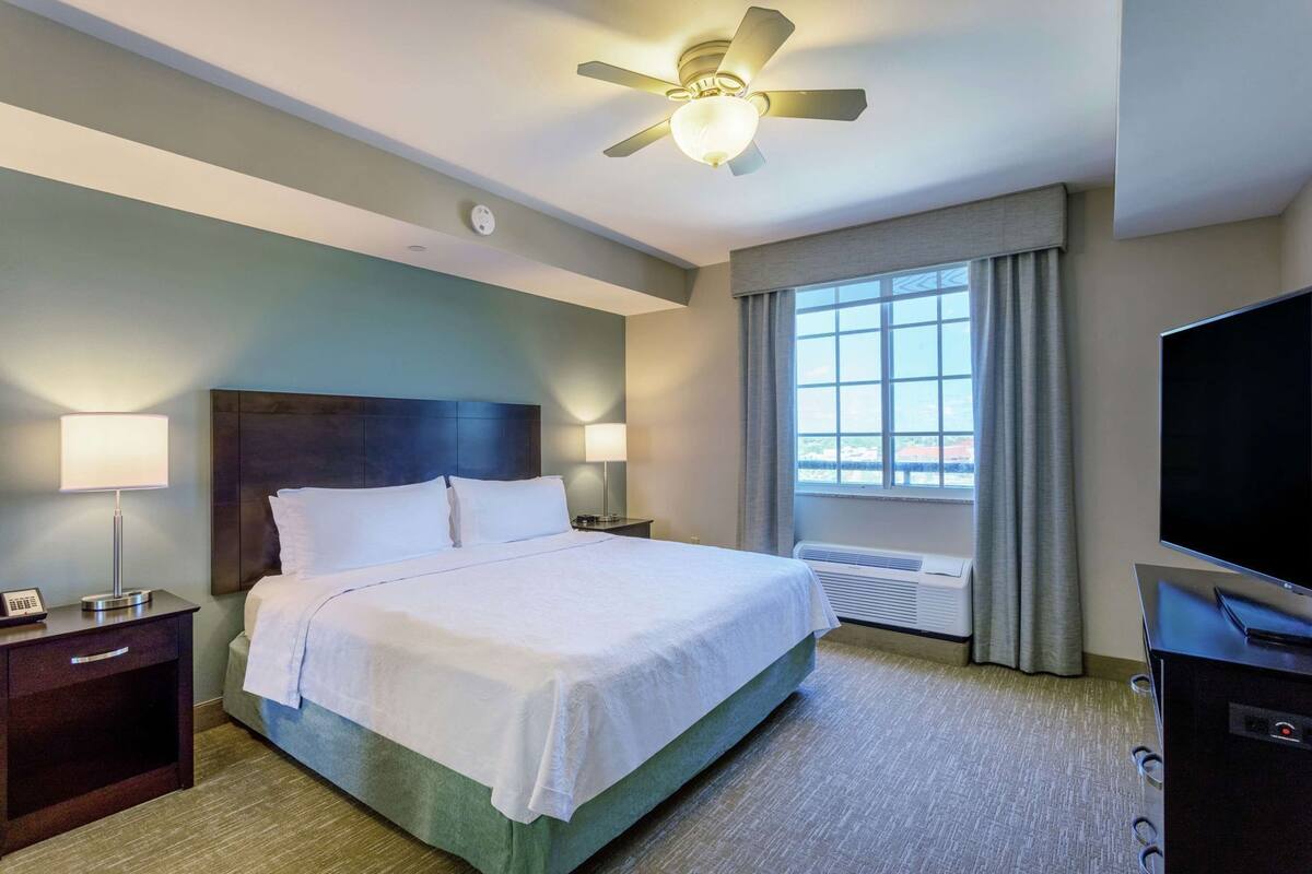 A hotel room with a bed, bedside lamps, a ceiling fan, a TV, and a window with curtains.