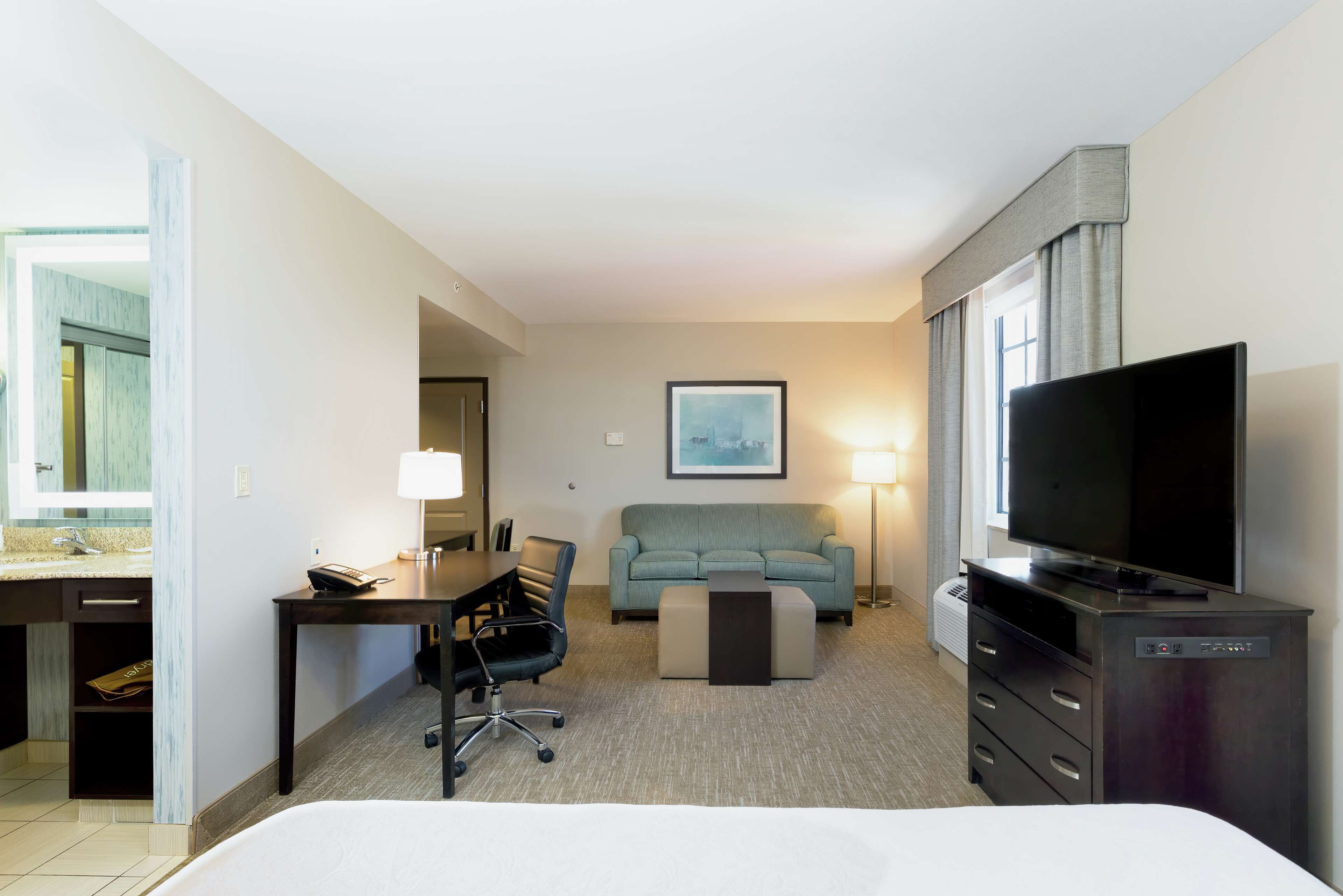 Habitación de hotel con cama, escritorio con silla, sofá, televisor y ventana con cortinas.