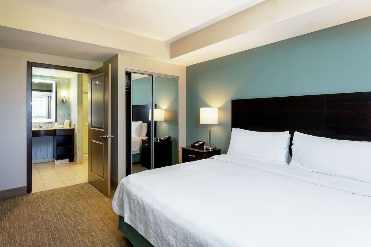 A hotel room with a large bed, a nightstand with a lamp, a mirror, and a view of the bathroom.