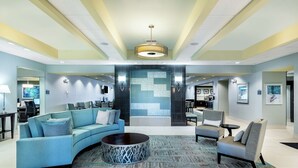 Un hall amplio con un sofá azul curvado, una mesa de centro redonda y un sillón gris. Una gran pantalla digital está montada en la pared, y hay dos lámparas de pared.