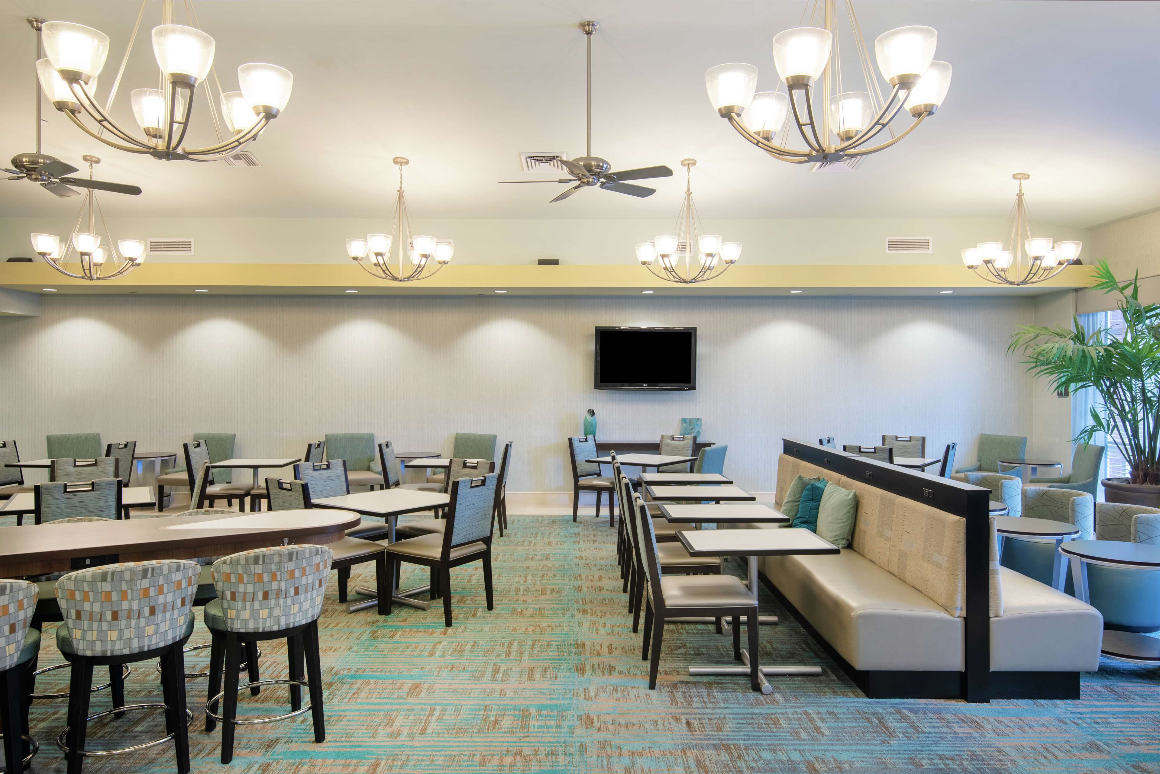 Espaço de refeições de hotel com várias mesas, cadeiras e um sofá. Há ventiladores de teto e lustres.