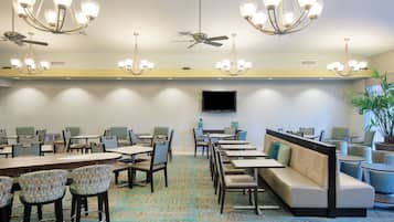 Espaço de refeições de hotel com várias mesas, cadeiras e um sofá. Há ventiladores de teto e lustres.