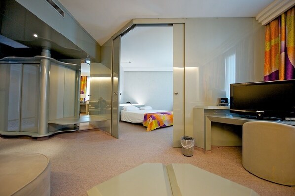 Habitación de hotel con cama, escritorio con televisor, sillón y minibar.