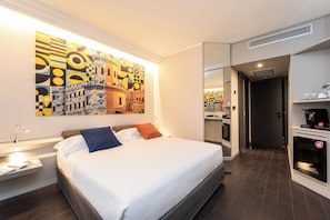 Habitación de hotel con una cama grande, una mesita de noche, un escritorio, una cafetera y una pared con un cuadro abstracto y colorido.